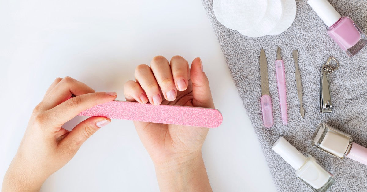 A hand holds a pink nail file to shape the nails on the other hand. Nail polish and clippers are off to the side on a cloth.